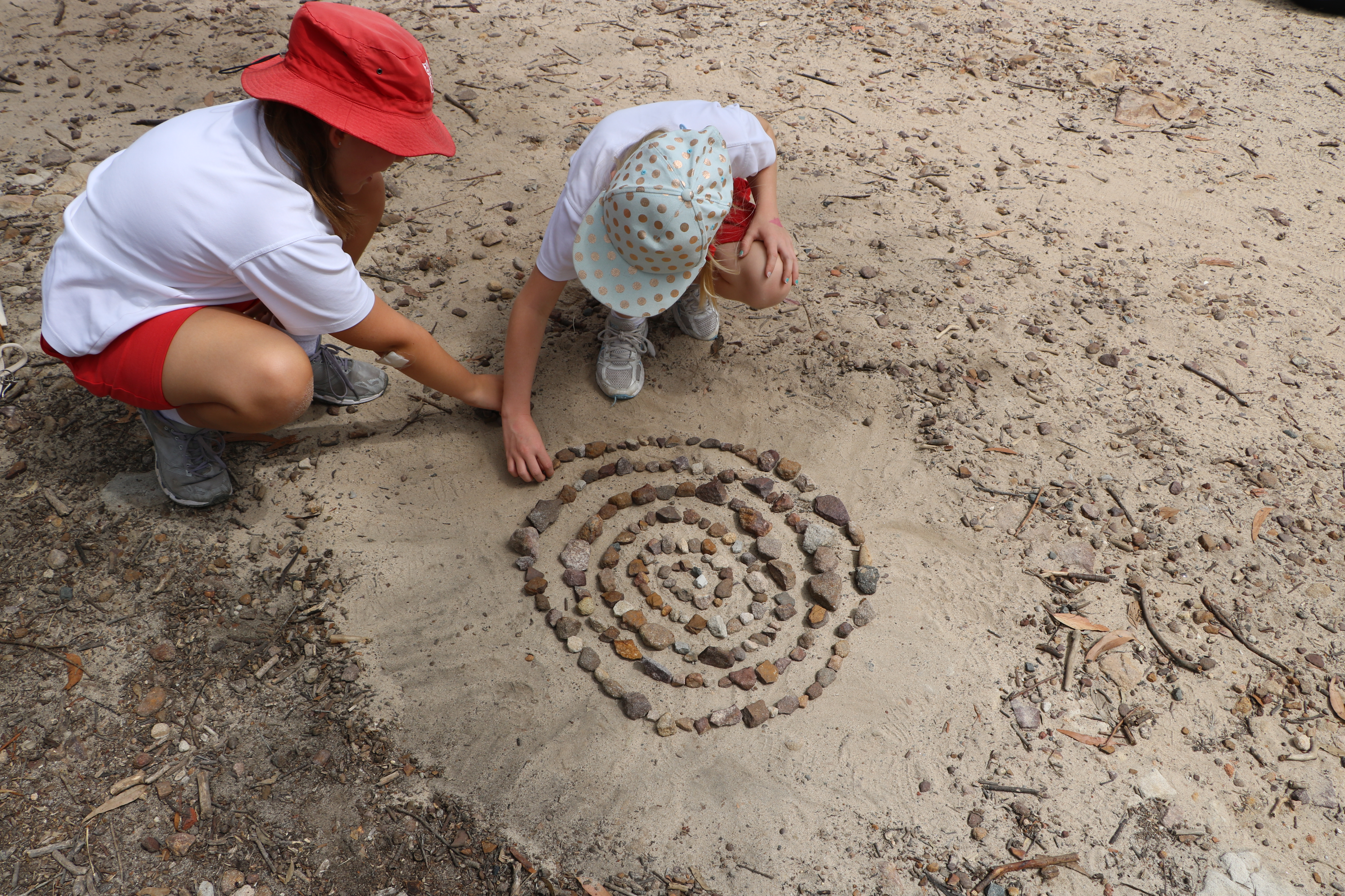 2 young students kneeling down in sand creating a spiral mandala with pebbles, rocks and twigs