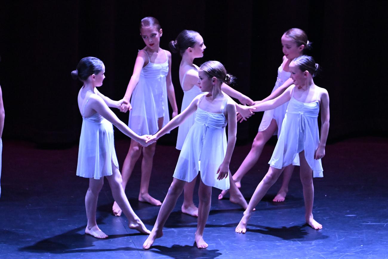 6 students in white dresses holding hands looking at each other on stage
