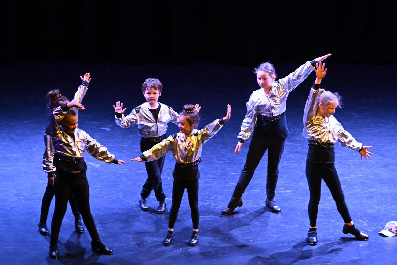 group of young students on stage in black pants and silver tops with black tap shoes