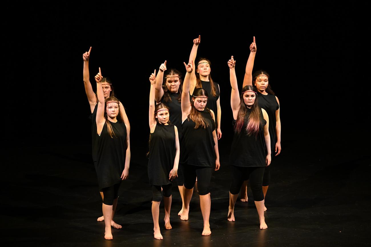 Students on stage in black dresses, one arm up in air
