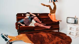 In a white room, an eclectically-dressed man in polka dots and ripped fish-net stockings on one end of a sofa, a topless person doing a headstand on the other end, and a person lying on the floor with a large round brown shaggy mat over their face and torso. An old TV on a crate.