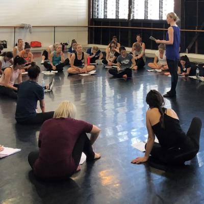 A large group of teachers sitting on a dance studio floor taking notes in front of a standing teacher.